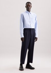 Camicia azzurro chiaro a quadri con colletto button-up, abbinata a pantaloni plissettati blu scuro e scarpe nere, su uno sfondo neutro.