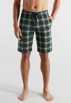 Mann trägt knielange Shorts in marineblauem und grünem Karomuster mit Kordelzugbund, steht barfuß mit einer Hand in der Tasche.