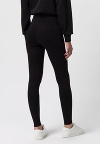 Černé legíny z hladkého, elastického materiálu, s vysokým pasem a přiléhavým střihem. Nosič má na sobě bílé tenisky.