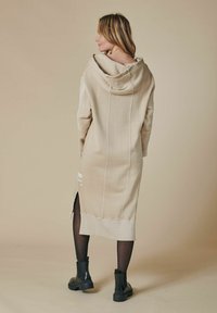 Langes beiges Kapuzenkleid mit Kapuze, seitlichen Schlitzen und weißen Nähten. Getragen mit schwarzen Knöchelstiefeln und transparenten Strumpfhosen.