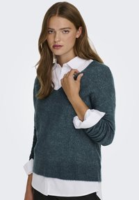 Türkiser Strickpullover mit V-Ausschnitt und geripptem Saum, über einem weißen Hemd mit Knopfleiste getragen. Hat lange Ärmel mit umgeschlagenen Bündchen.