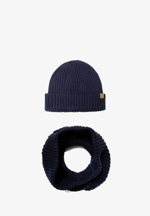Bonnet tricoté bleu marine avec un motif côtelé et une étiquette en cuir, associé à une écharpe infinity assortie présentant une texture côtelée similaire.