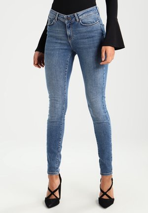 Slim-fit blauwe spijkerbroek gedragen met een zwarte top met lange mouwen en zwarte hoge hakken met gekruiste bandjes.