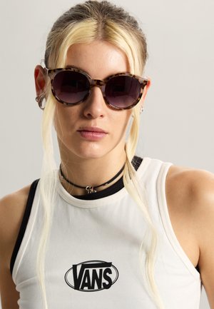 Joven con gafas de sol con montura carey, una camiseta sin mangas blanca de Vans y un collar negro tipo gargantilla, sobre un fondo liso.