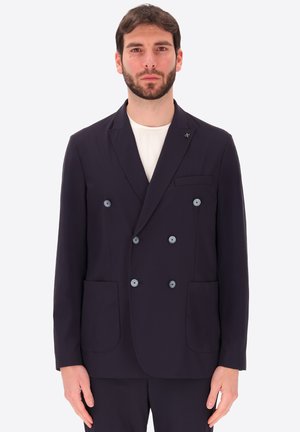Uomo con barba che indossa un blazer doppiopetto scuro e una camicia bianca, in piedi davanti a uno sfondo chiaro uniforme.