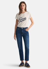Helle T-Shirt mit blauem "Gant"-Logo, kombiniert mit dunkelblauen Straight-Leg-Jeans. Das Model trägt blaue Sneakers und hat die Hände in den Taschen.
