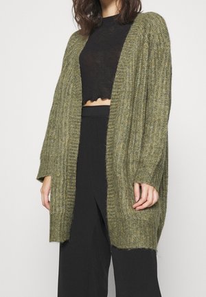 Kvinde iført en olivengrøn ribstrikket cardigan over en sort cropped top og sorte højttaljede bukser, stående mod en ensfarvet baggrund.