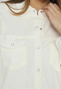 Blouse blanche boutonnée avec deux poches poitrine, fermetures pression et un col. Le tissu semble lisse et léger.