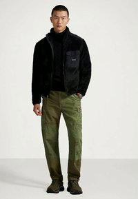 Man wearing black zip-up fleece jacket, black turtleneck, patchwork green cargo pants, and dark sneakers, standing against plain light background.