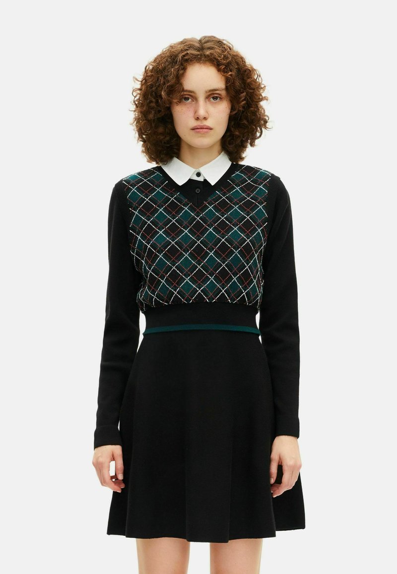 Jovem mulher com cabelo encaracolado, vestindo um vestido preto de mangas longas, um colete em padrão argyle verde e branco e uma gola branca.