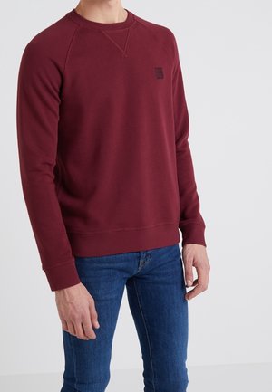Burgundy sweatshirt med rund halsringning, ribbad mudd och nederkant. Har en liten svart logotyp på bröstet. Kombinerad med blå jeans.