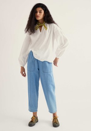 Frau mit lockigem Haar, die eine weiße Bluse mit langen Ärmeln, hellblaue, weit geschnittene Hosen, olivgrüne Sneaker und ein olivgrünes Halstuch trägt.