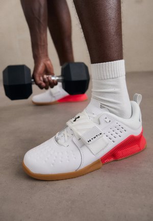 Vita träningsskor med en texturerad yta, grå accenter, röd häl och en gummisula. Har ett rem för säker passform runt mellanfoten.