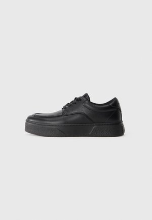 Zapatilla negra de cuero con cordones, suela gruesa y texturizada, punta redondeada y altura baja en el tobillo sobre un fondo blanco liso.