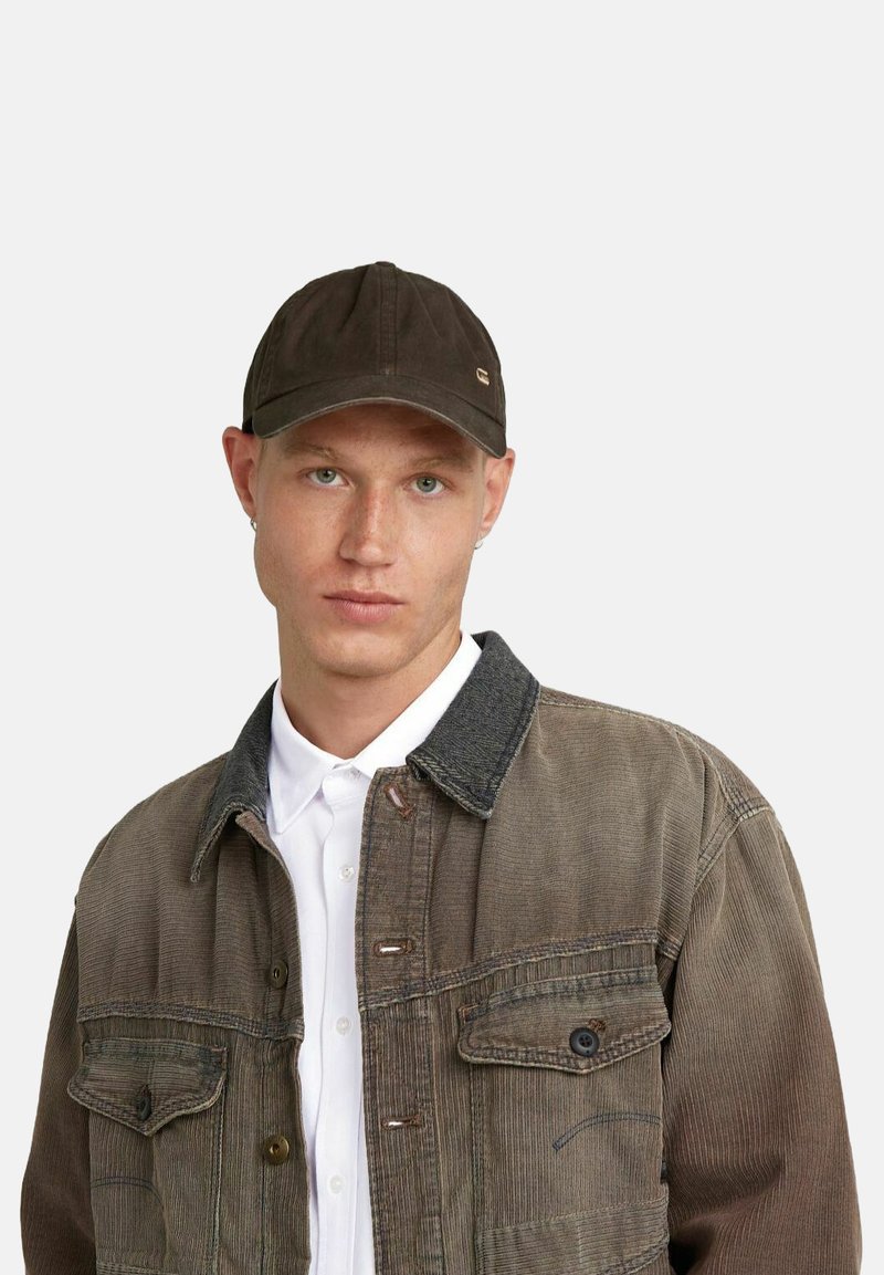 Cappello marrone in twill di cotone; design a sei pannelli con visiera curva e dettagli metallici. Indossato con una giacca di velluto a coste beige e una camicia bianca.