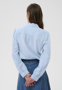 Chemise en coton bleu clair avec un col à volants, des manches longues bouffantes et un détail froncé au dos, associée à une jupe en denim.