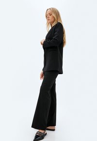 Zwarte op maat gemaakte blazer met notch-lapels, gecombineerd met flared zwarte broek. Het model draagt pointed zwarte schoenen met hardware accenten.