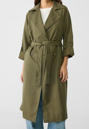 Femme portant un trench-coat ceinturé vert olive avec les manches retroussées, par-dessus un haut blanc et un jean bleu, sur un fond uni.