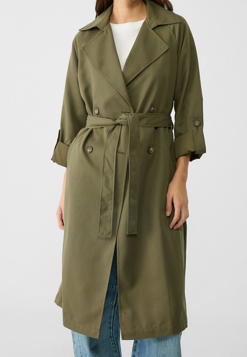 Femme portant un trench-coat ceinturé vert olive avec les manches retroussées, par-dessus un haut blanc et un jean bleu, sur un fond uni.