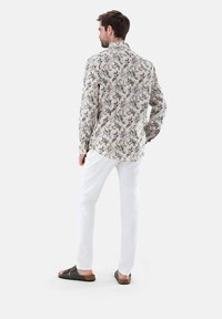 Camicia a maniche lunghe con fantasia floreale in tessuto leggero, caratterizzata da tonalità di verde e beige, indossata con pantaloni bianchi e sandali scuri.