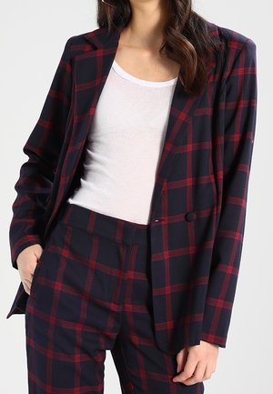 Vrouw in een marineblauw pak met een rood geruit patroon en een witte top met een ronde hals, staand met één hand in haar zak.