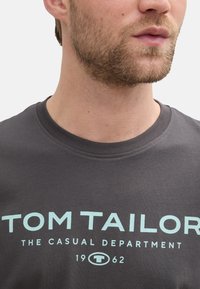 Tricou gri din bumbac, cu guler rotund. Conține un text de culoare albastru deschis "TOM TAILOR THE CASUAL DEPARTMENT 1962" afisat proeminent.
