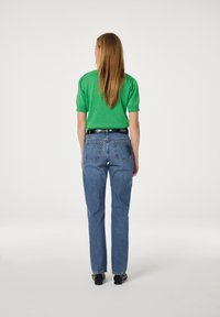 Vrouw met lang haar, dressed in een groene top met korte mouwen, blauwe high-waisted jeans, een zwarte riem en zwarte schoenen, staande met haar rug naar de kijker gekeerd.