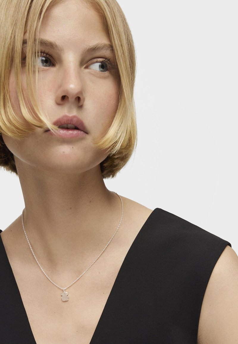 Collar de plata con un charm redondo, llevado por una persona con una blusa negra. El charm presenta una superficie lisa y reflectante, diseño de cadena sencillo.