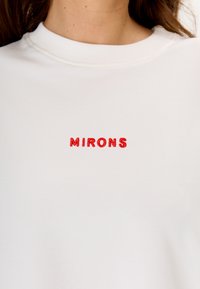 Witte sweatshirt met rode geborduurde tekst "MIRONS" aan de voorkant. Ronde hals en een zachte, gladde stofstructuur.