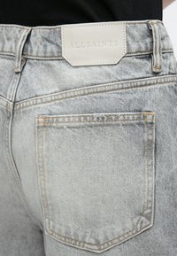 Heledast hallid teksapüksid, millel on tagatasku, oranžid õmblused ja tekstuuriga valgest nahast silt, millele on sissepressitud brändinimi "ALLSAINTS".