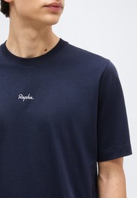 Νεαρό άτομο που φοράει μπλε σκούρο μπλουζάκι Rapha, ορατό από τον λαιμό έως το μέσο του κορμού, μπροστά σε ένα απλό ανοιχτόχρωμο φόντο.