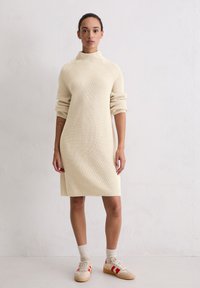 Kremowa dzianinowa sukienka z wysokim kołnierzem, żebrowaną fakturą i opuszczonymi ramionami. Do tego beżowe trampki z czerwonymi paskami i białymi akcentami na skarpetach.