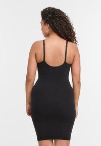 Donna con capelli ricci che indossa un abito aderente nero senza maniche con spalline regolabili, vista di spalle su uno sfondo bianco.