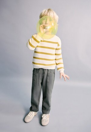 Bambino piccolo con maglione a righe e pantaloni grigi che soffia una grande bolla gialla contro uno sfondo neutro.