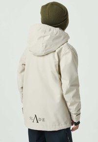Beige wasserdichte Jacke mit Kapuze, ausgestattet mit verstellbaren Manschetten und einem Logo auf der Rückseite. Glatte Textur, hüftlange Schnittform.
