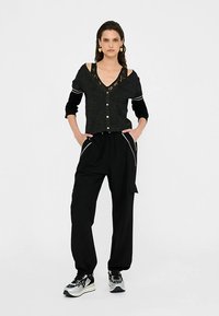 Svart kofta med spetsdetaljer över en svart topp, kombinerad med svarta byxor med vita kantband och svarta och grå sneakers.