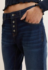 Primer plano de un pantalón de mezclilla azul oscuro con cuatro botones de metal, costuras naranjas y una mano parcialmente dentro del bolsillo delantero.