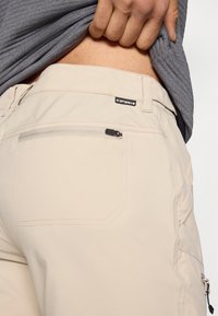 Μπεζ εξωτερικό παντελόνι με φερμουάρ σε τσέπη στο πίσω μέρος, κατασκευασμένο από ελαφρύ ύφασμα. Διαθέτει ετικέτα μάρκας κοντά στη μέση.