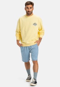 Yellow cotton sweatshirt with a front logo, paired with light blue denim shorts. Black sneakers and grey socks complete the outfit.