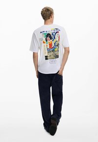 Valge T-särk, mille seljal on graafiline print, mis sisaldab Jaapani kultuuri elemente, kombineeritud tumesiniste pükste ja mustade jalanõudega.