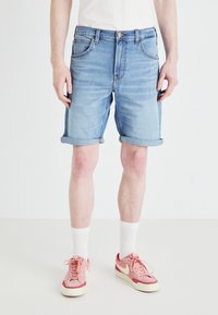 Ανοιχτό μπλε τζιν σορτς με διπλωμένα στρινγκ, πέντε τσέπες και κούμπωμα με κουμπί. Φοριέται με ροζ αθλητικά παπούτσια και λευκές κάλτσες.