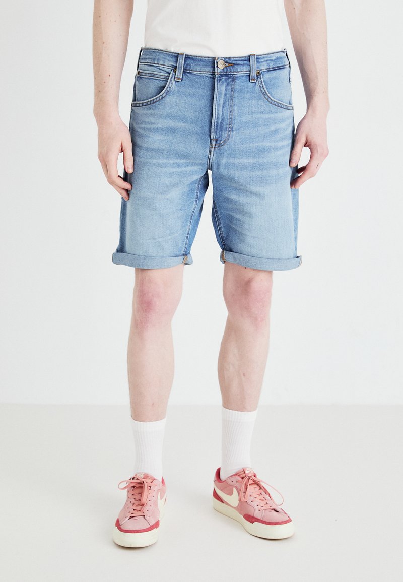 Ανοιχτό μπλε τζιν σορτς με διπλωμένα στρινγκ, πέντε τσέπες και κούμπωμα με κουμπί. Φοριέται με ροζ αθλητικά παπούτσια και λευκές κάλτσες.