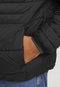 Schwarze Puffy-Jacke mit strukturierter Oberfläche, die eine Hand in der Tasche zeigt. Verfügt über elastische Bündchen und horizontale Steppnähte.