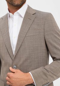 Jasnobrązowy marynarka skrojona na miarę z teksturowanej tkaniny, zapinana na jeden guzik, z klapami w kształcie notch. Biała koszula wystaje spod spodu.