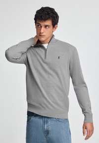 Grauer Pullover mit halbem Reißverschlusskragen und gerippten Bündchen. Hergestellt aus weichem Stoff, mit einem kleinen Logo auf der linken Brust. Getragen mit hellen Jeans.