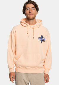 Sudadera color melocotón con un bolsillo canguro, capucha con cordón y un logotipo en forma de diamante negro y azul a la izquierda. Tejido suave, corte relajado.