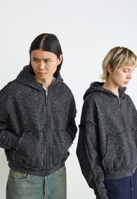 Black zip-up hoodies with a cracked pattern, distressed edges, and front pockets. Loose fit with a textured finish.