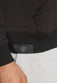 Tessuto in felpa nera con orlo a costine. Presenta una tasca laterale e un'etichetta con logo tono su tono. Abbinato a pantaloni grigi.