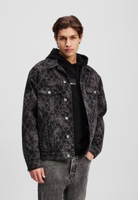 Giacca di jeans nera con un motivo geometrico, due tasche sul petto, bottoni in metallo, indossata sopra un hoodie nero.