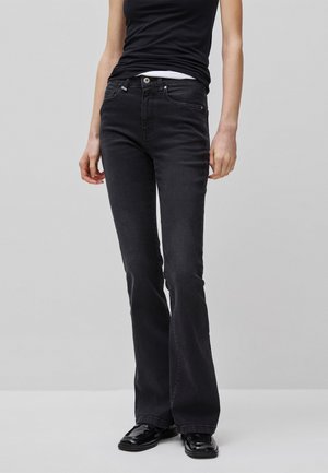 Persona che indossa jeans neri a zampa, top nero senza maniche e scarpe nere lucide con punta quadrata, in piedi davanti a uno sfondo chiaro e semplice.